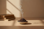 Smoldering incense stick in a bowl on a wooden surface with books in the background